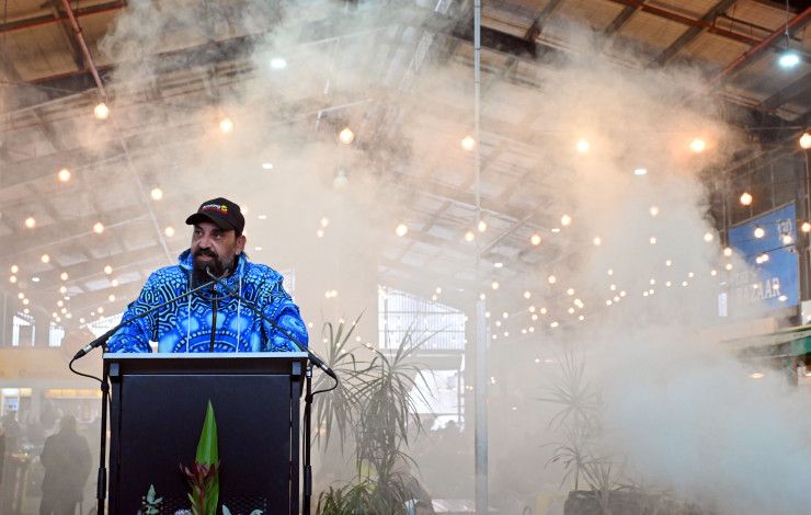 A man stands behind a lectern as smoke creates and almost white background behind him.
