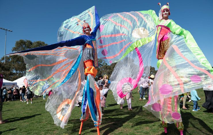 Two characters on stilts with flowing fairy wings walking through a park on a blue sky day.