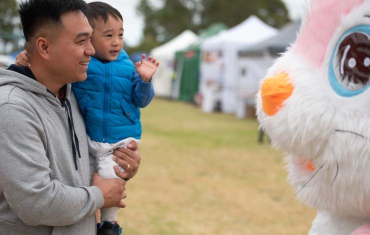 A man holds a small child up while they look into the eyes of a giant pink bunny