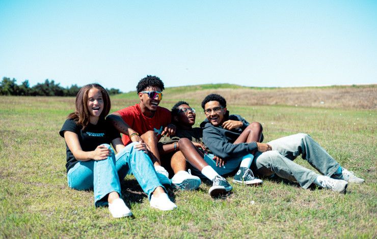 Four young people sitting together and laughing on a grassy hill.