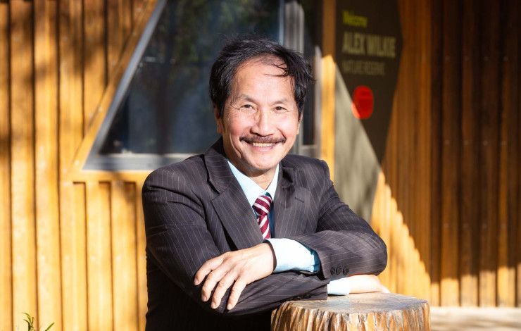 A man with dark hair and a moustache smiles at the camera with his arms crossed in front of him