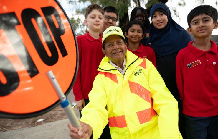 A smiling man dressed in high vis holding a stop sign while surrounded by smiling school children