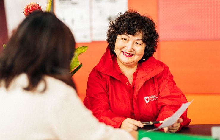 A woman dressed in a bright red shirt sits at a table and explains something to a woman sat opposite her with her back to the camera.