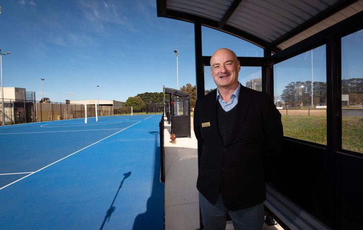 A man stands under a shelter next to a blue netball court.