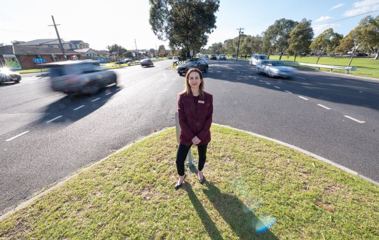 A woman dressed in a suit standing on a grass island in the middle of a busy road.