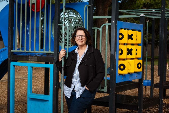 A woman wearing a dark jacket and striped shirt leaning against children's play equipment while smiling at the camera.
