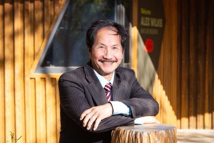 A man with dark hair and a moustache smiles at the camera with his arms crossed in front of him