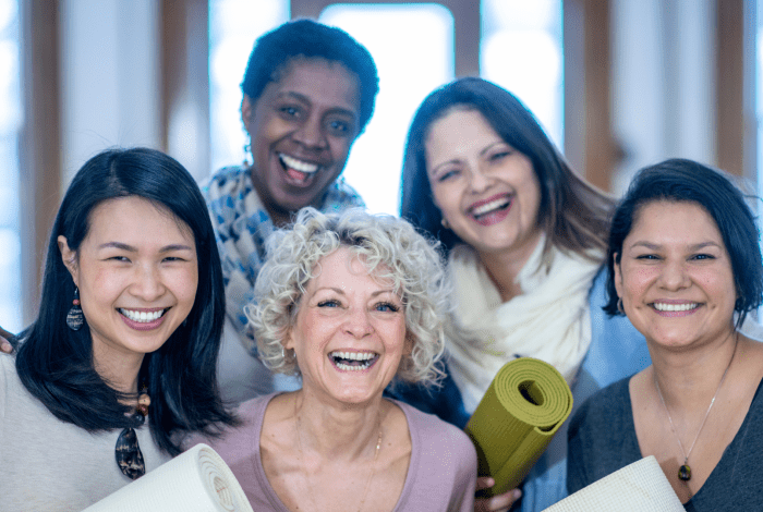 Five people of different ages and cultural backgrounds are standing together in a group. They are smiling and holding yoga mats.