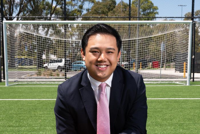 A man in a dark suit crouches on one knee on a synthetic soccer pitch while holding an orange soccer ball.