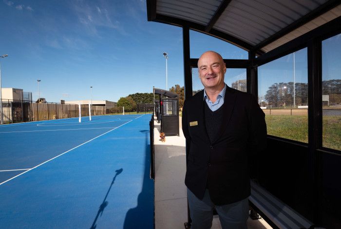 A man stands under a shelter next to a blue netball court.