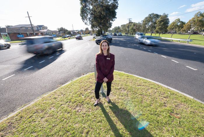 A woman dressed in a suit standing on a grass island in the middle of a busy road.