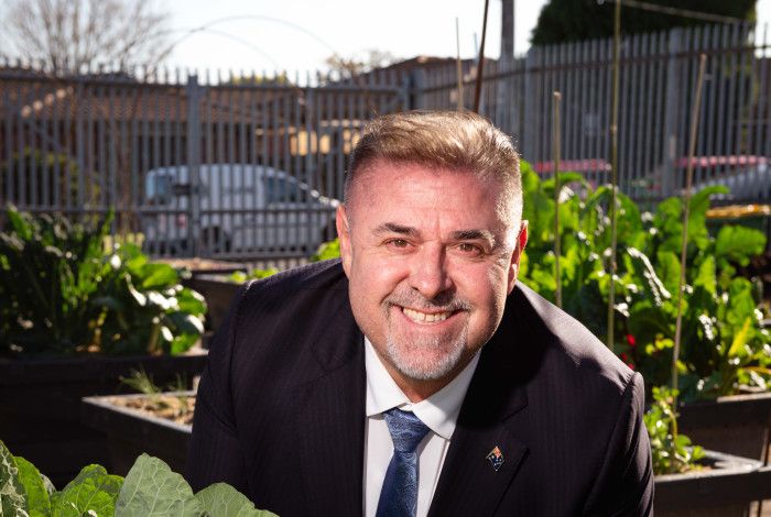 Man smiling at the camera crouching behind a leafy green plant.