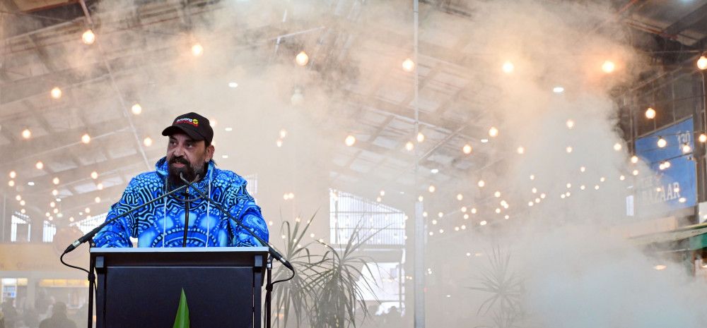 A man stands behind a lectern as smoke creates and almost white background behind him.