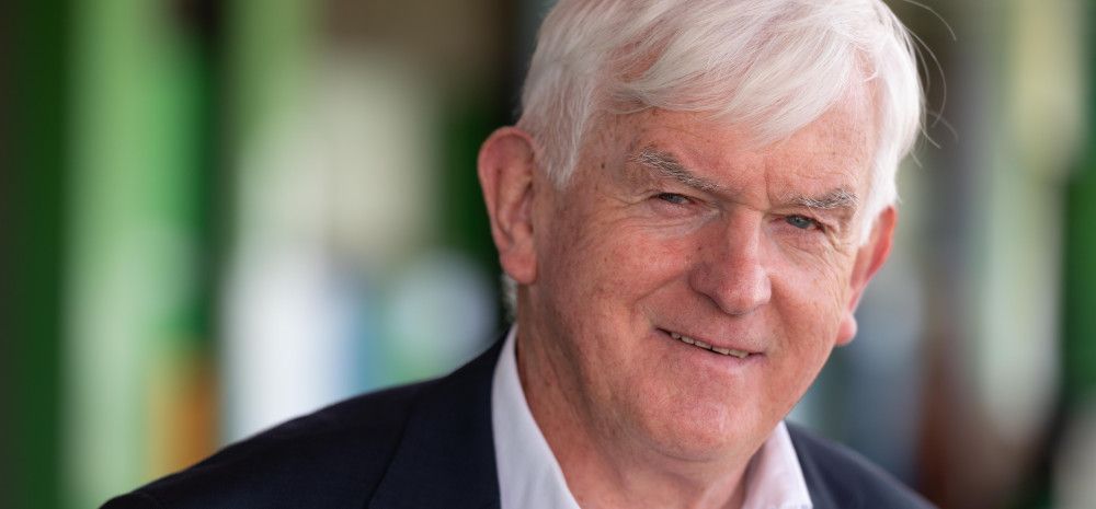 Man with white hair, dressed in a white shirt and black jacket, smiling at the camera