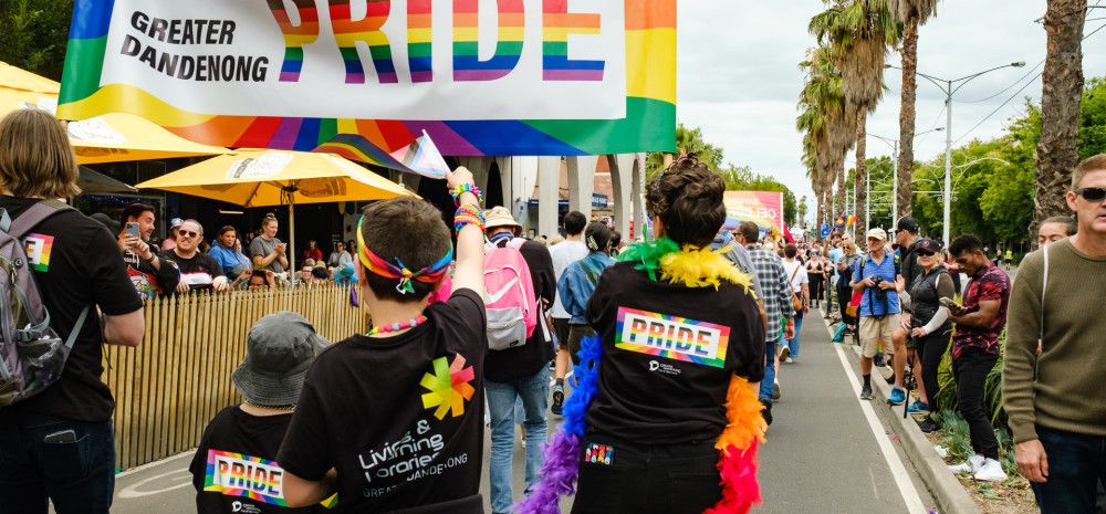 A group of people walking down a street lined by crowds under a banner that reads Greater Dandenong Pride
