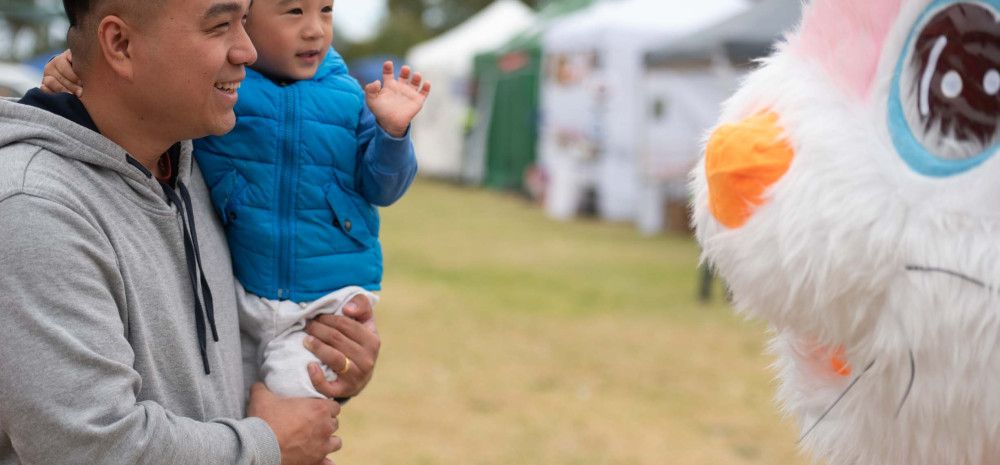A man holds a small child up while they look into the eyes of a giant pink bunny
