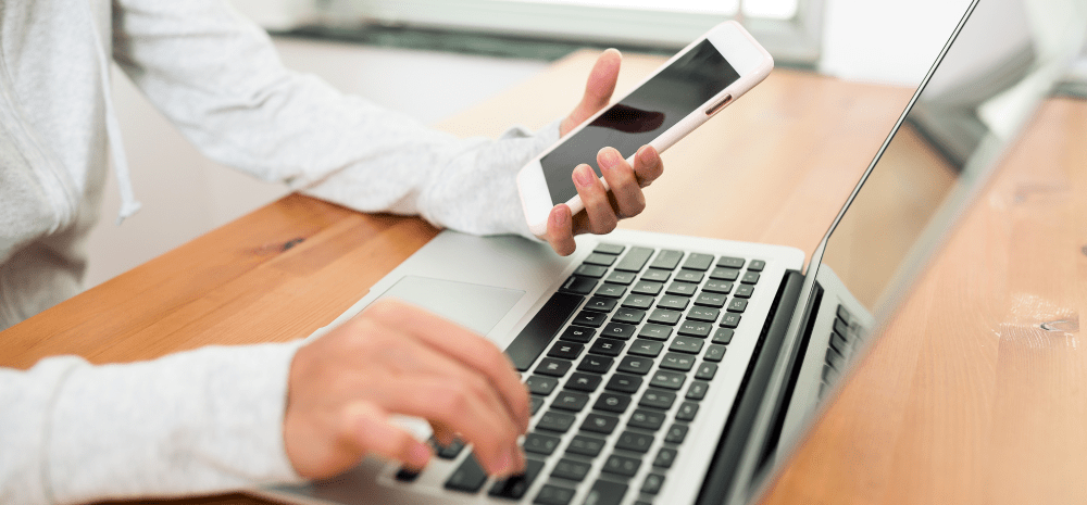 A person holding a smart phone and typing on a laptop with the other hand.