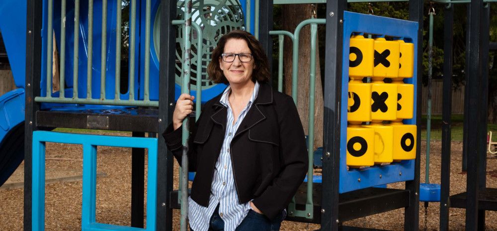 A woman wearing a dark jacket and striped shirt leaning against children's play equipment while smiling at the camera.