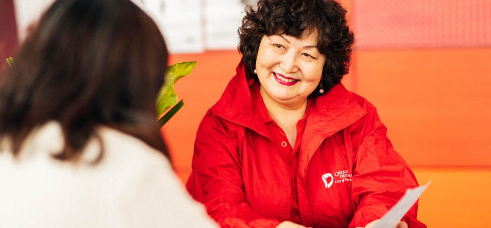 A woman dressed in a bright red shirt sits at a table and explains something to a woman sat opposite her with her back to the camera.