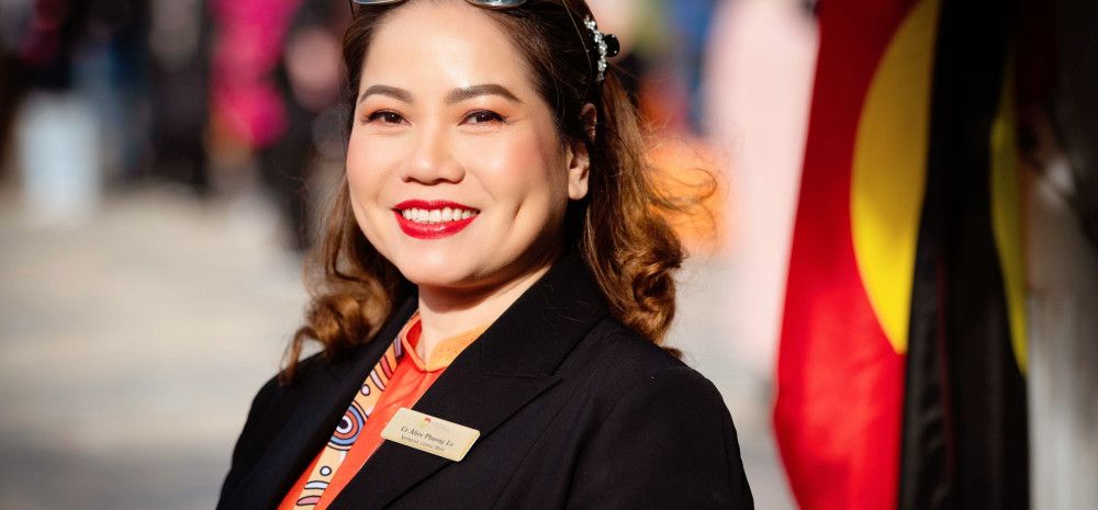A smiling woman in a black jacket stands side on and smiles to the camera with an Aboriginal flag in the background.