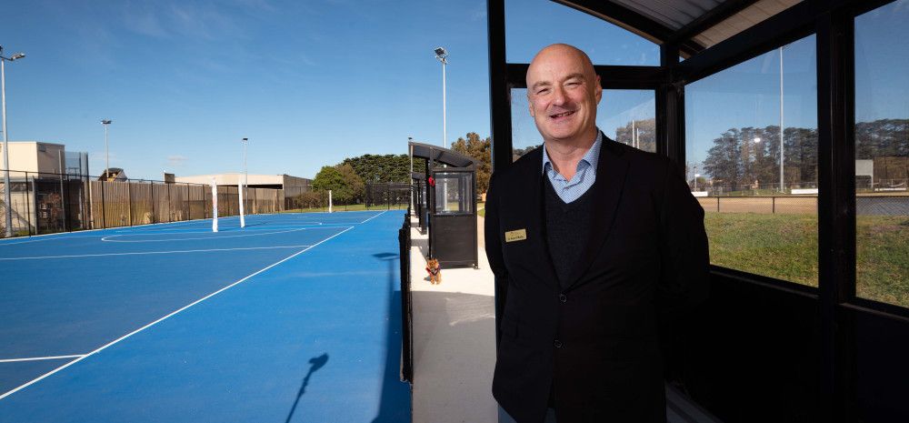 A man stands under a shelter next to a blue netball court.