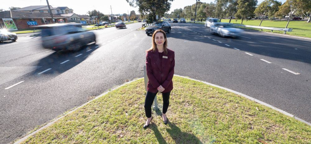 A woman dressed in a suit standing on a grass island in the middle of a busy road.