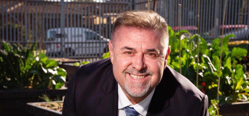 Man smiling at the camera crouching behind a leafy green plant.
