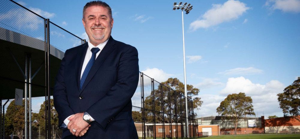 Man in a dark suit stands on a sports ground under a large light tower