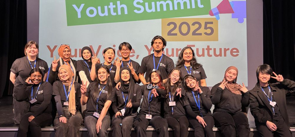 A large group of young people pose together on a stage in front of a sign that reads Youth Summit 2025