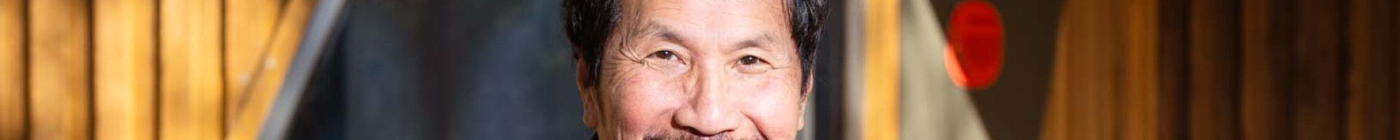 A man with dark hair and a moustache smiles at the camera with his arms crossed in front of him