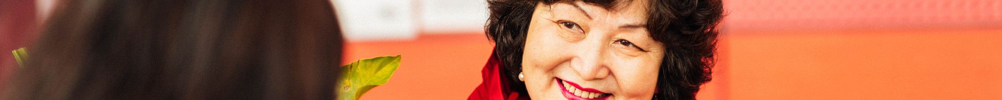 A smiling woman dressed in a bright red shirt sits at a table and explains something to a woman sat opposite her with her back to the camera.