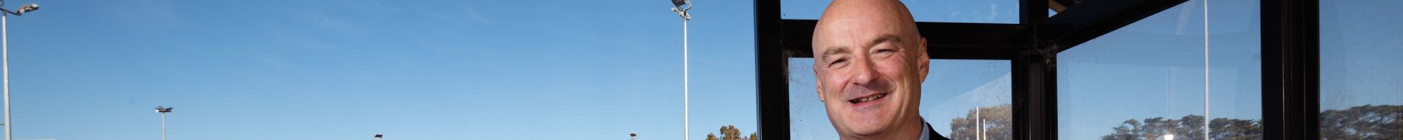A man stands under a shelter next to a blue netball court.