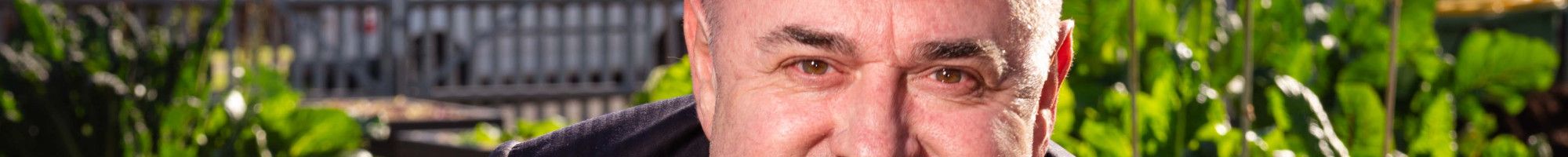 Man smiling at the camera crouching behind a leafy green plant.